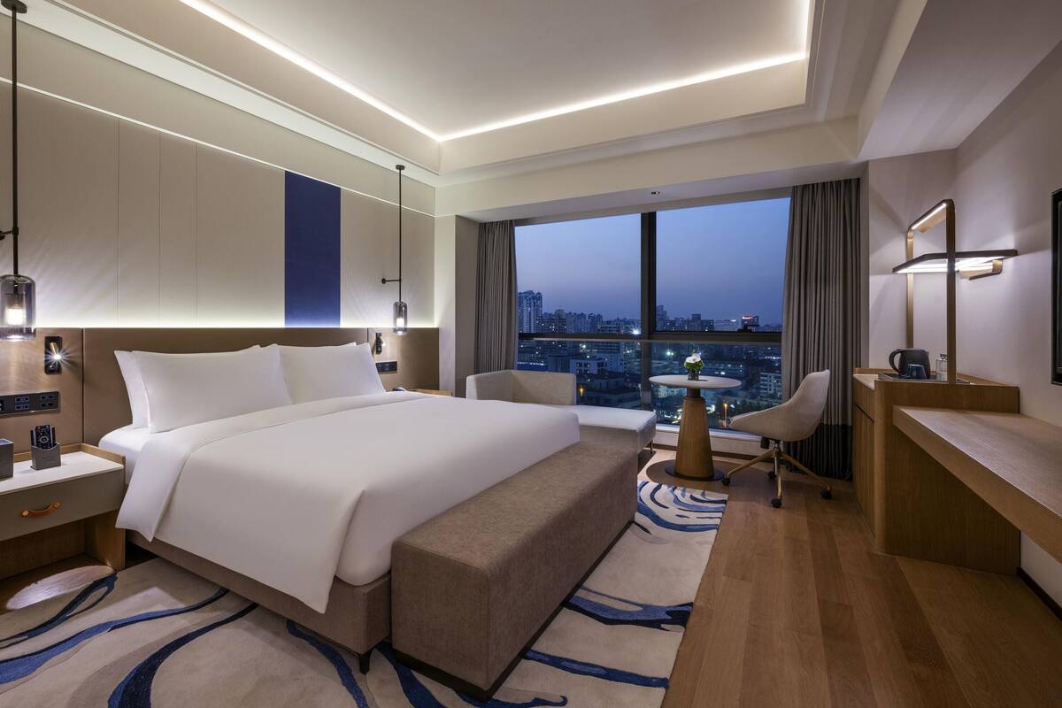 A modern hotel room with a large bed, a desk, a chair, and a view of the cityscape.