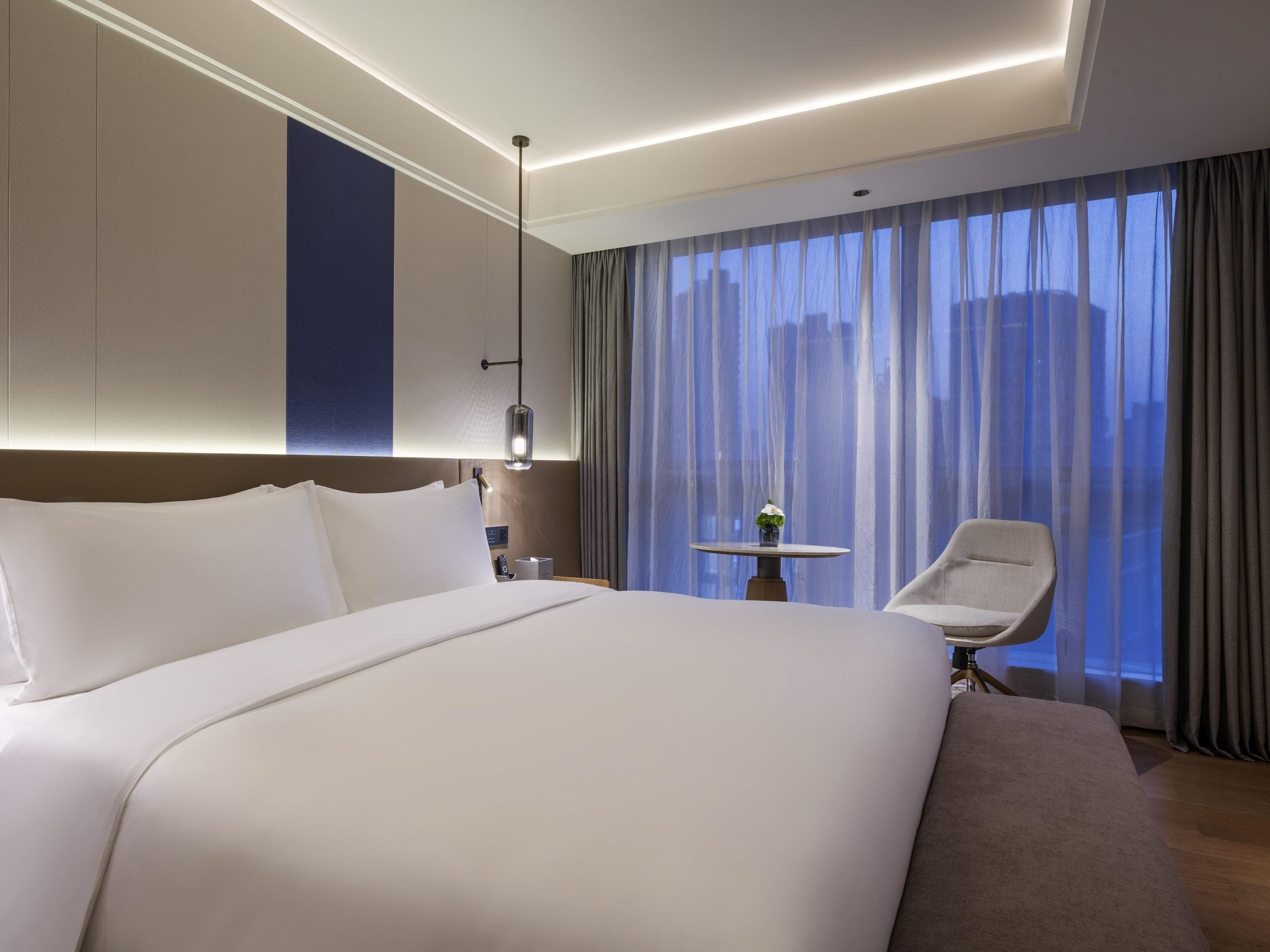 Moderni hotellihuone, jossa on suuri sänky, pieni pöytä maljakolla, tuoli ja näkymä rakennuksiin ohuiden verhojen läpi.