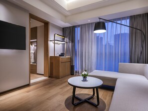 A modern hotel room with a flat-screen TV, a round table with a vase, a sofa, a desk with a lamp, and a view of the city through large windows. - Novotel Shanghai Jingan (Shanghai)