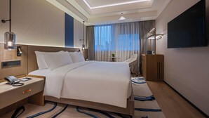 A modern hotel room with a large bed, bedside table with a phone, a desk with a lamp, a TV mounted on the wall, and a view of the city through the window. - Novotel Shanghai Jingan (Shanghai)