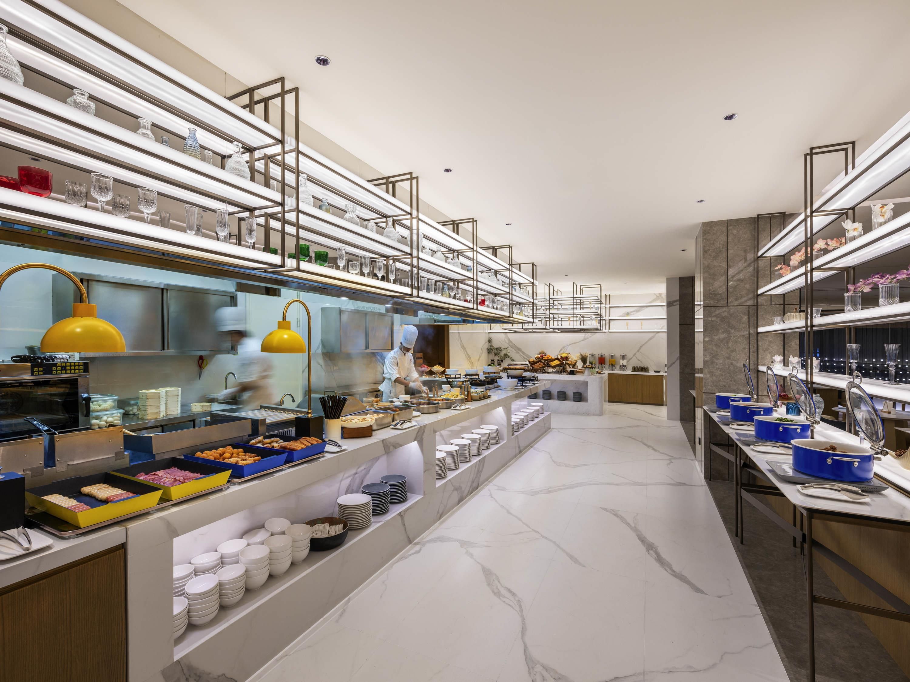 Un buffet moderne, avec plusieurs présentoirs de plats, de la verrerie et un chef qui prépare les aliments.