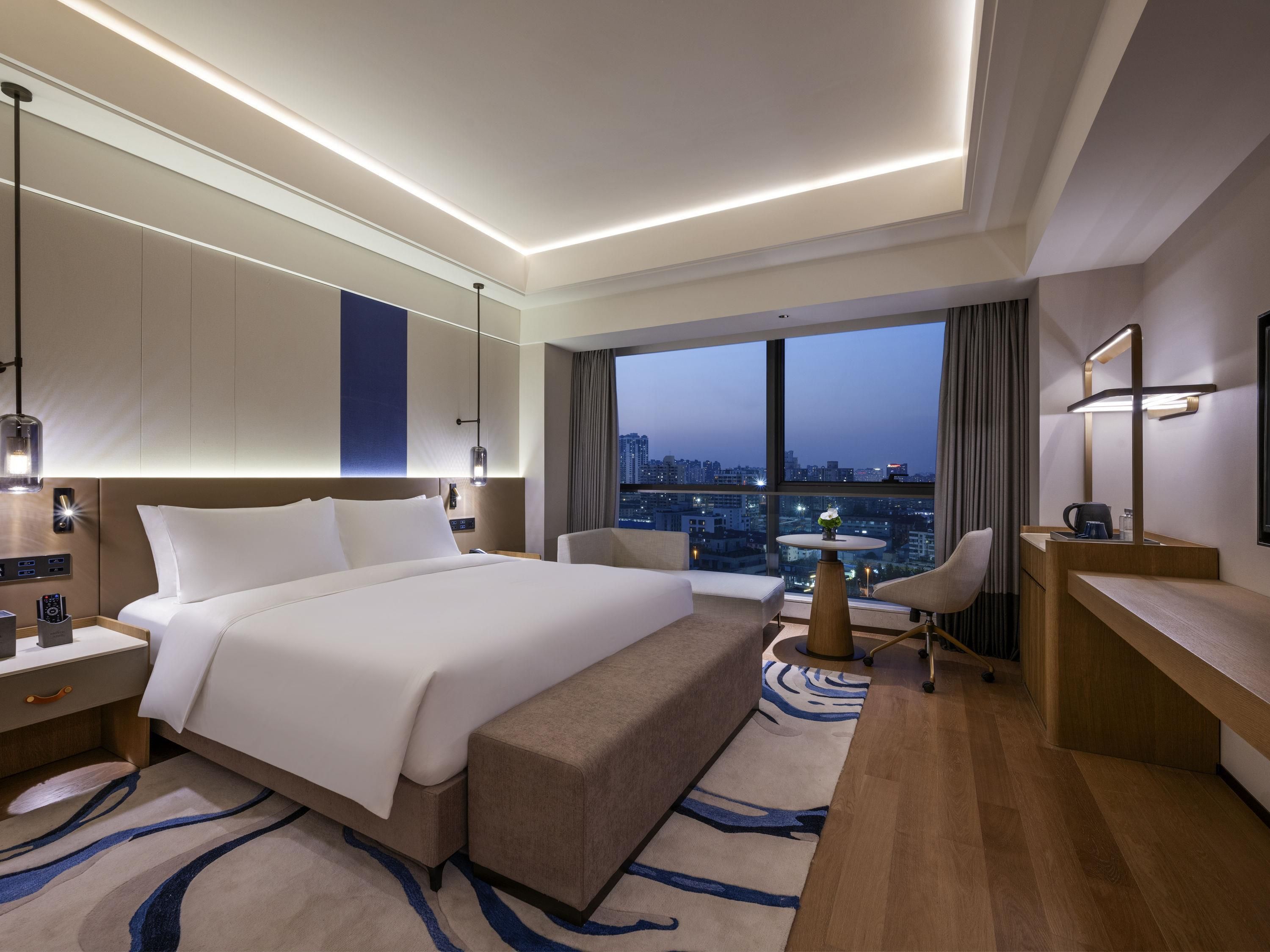 Habitación de hotel moderna con una cama grande, un escritorio, una silla y vistas a la ciudad.