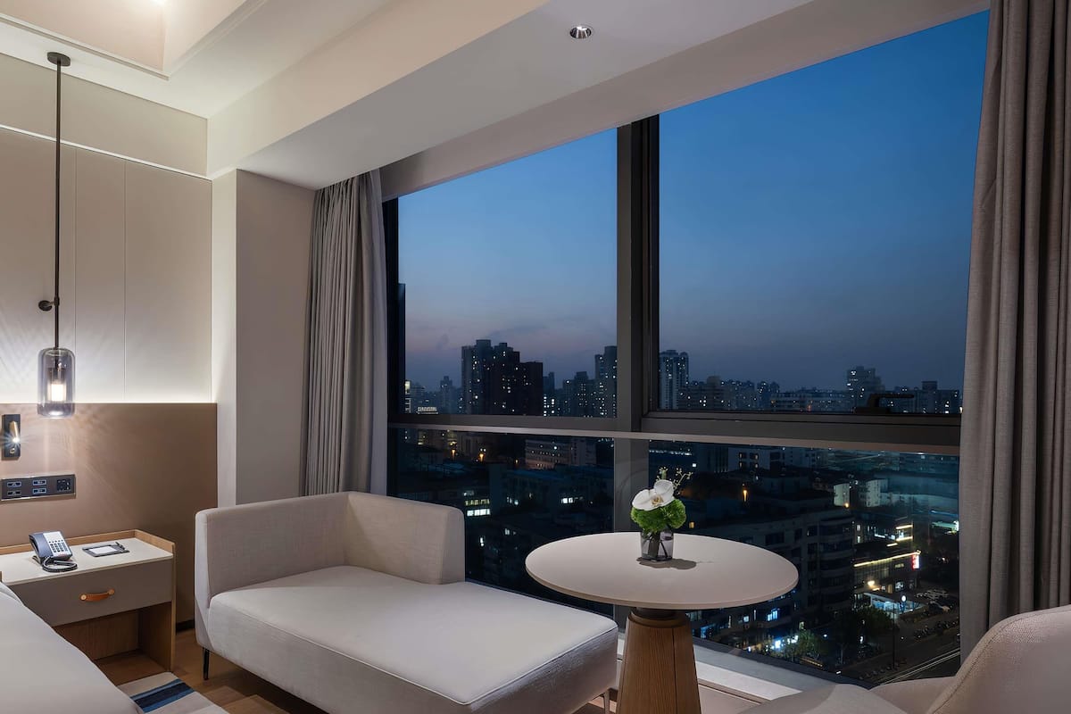 A modern hotel room with a city view, a sofa, a small table, and a nightstand with a phone and charger.