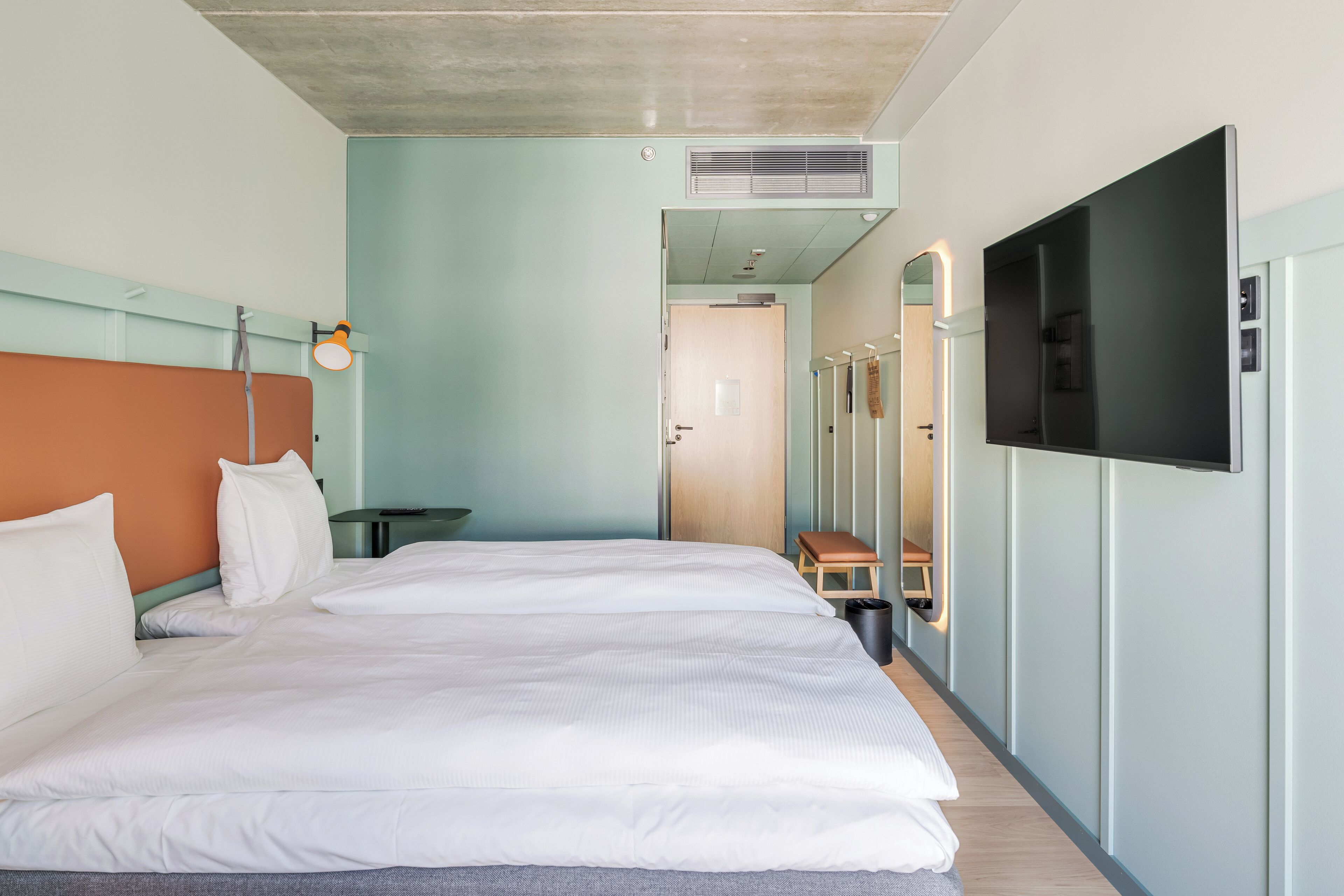 A modern hotel room with a large bed, a TV mounted on the wall, and a small table with a lamp.