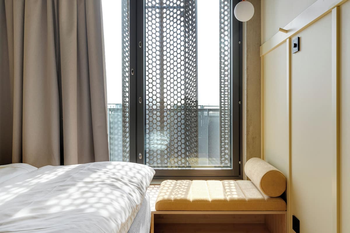 Un dormitorio con cama, ventana con vista a un edificio y una banca con cojín.