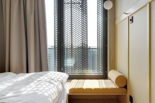 A bedroom with a bed, a window with a view of a building, and a bench with a cushion.