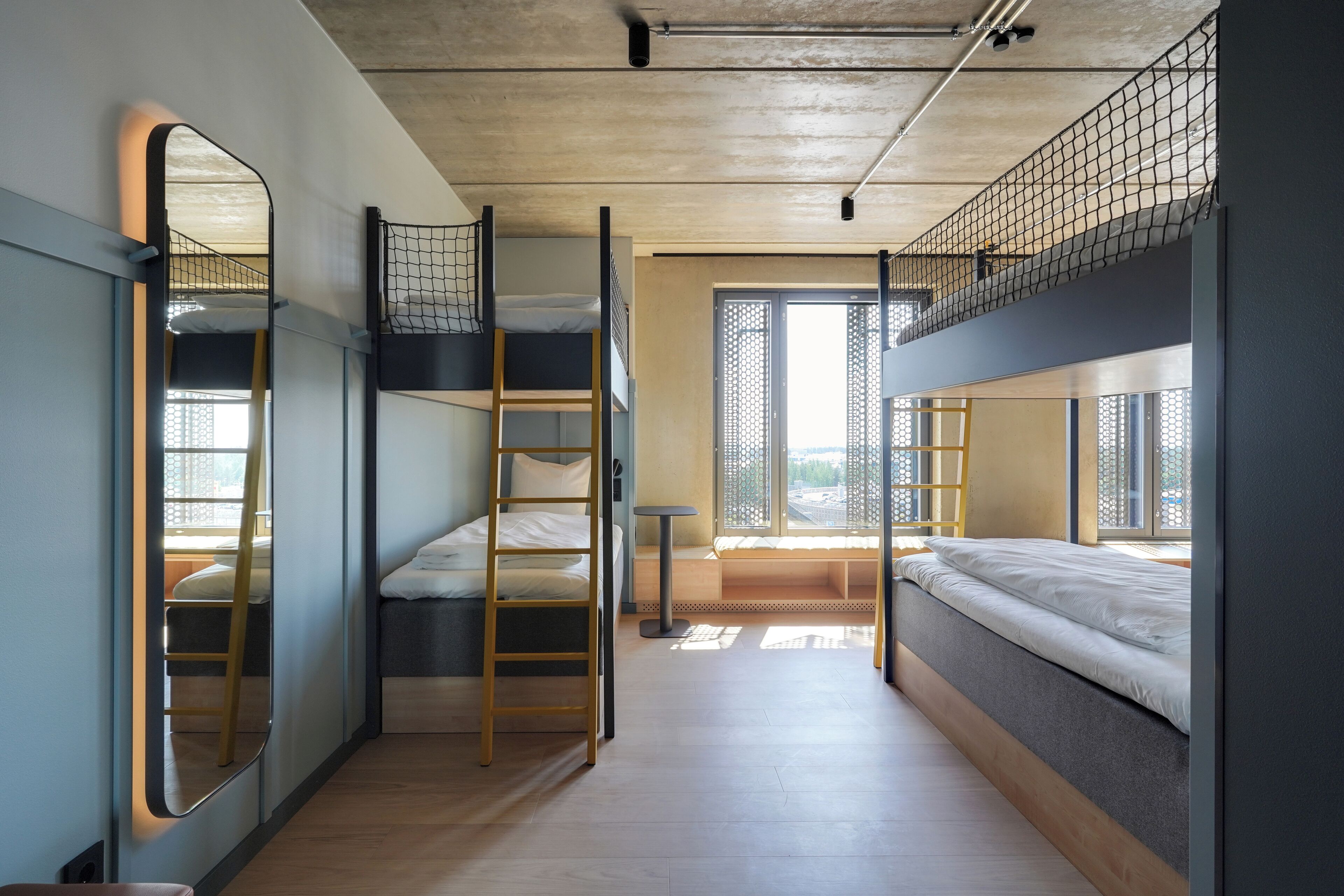 A dorm-style room with bunk beds, a ladder, a small table, and a window with a view.