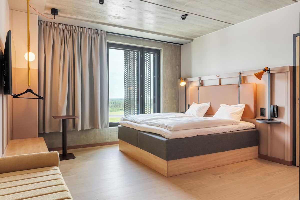 Una habitación de hotel moderna con una cama grande, una mesita de noche, un televisor y una ventana con cortinas.