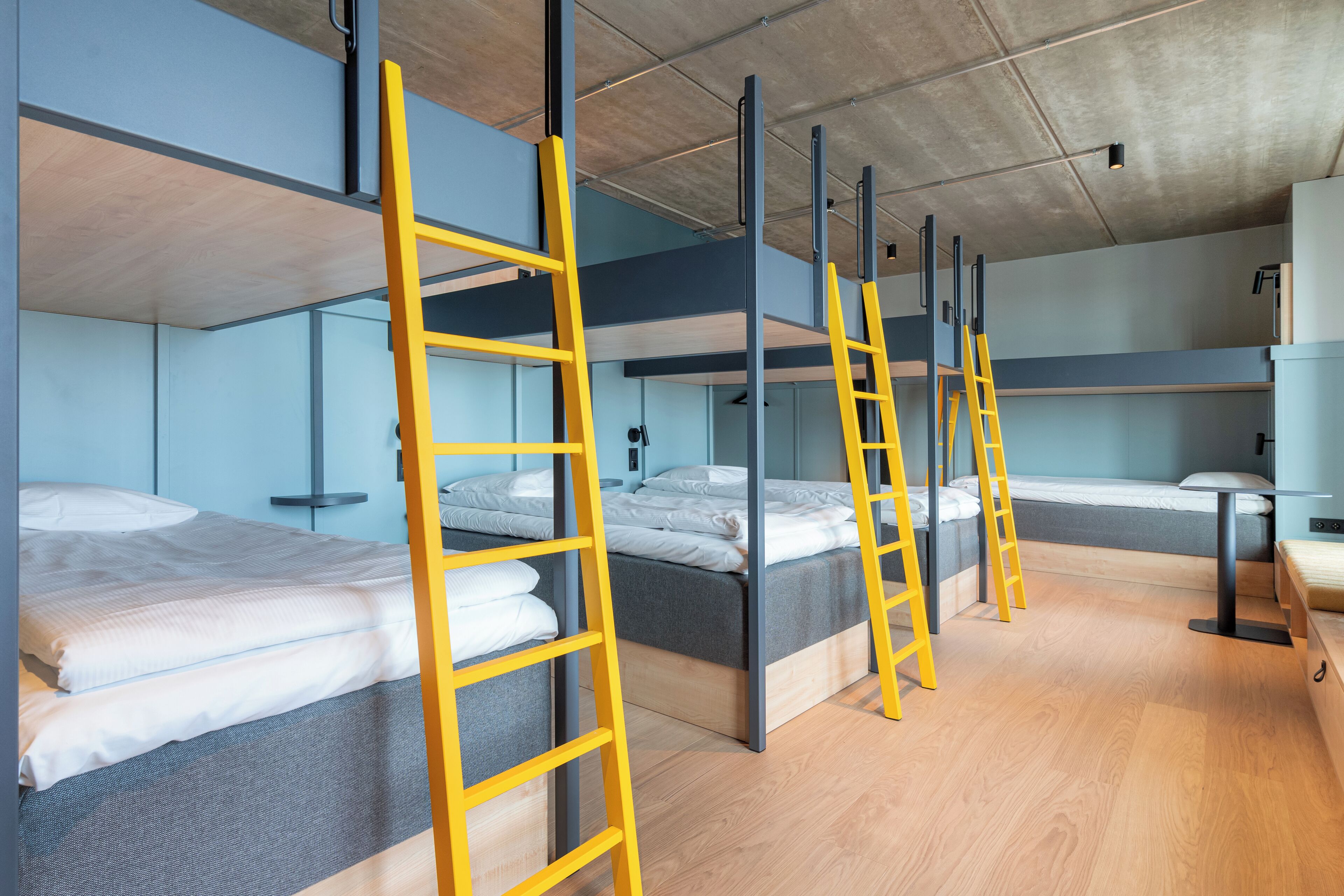 Quarto em dormitório com beliches, piso de madeira e teto de concreto.
