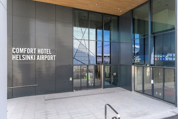 L’extérieur du Comfort Hotel, situé à l’aéroport d’Helsinki, se distingue par ses grandes portes vitrées, sa façade moderne et le bâtiment du terminal de l’aéroport visible en arrière-plan.