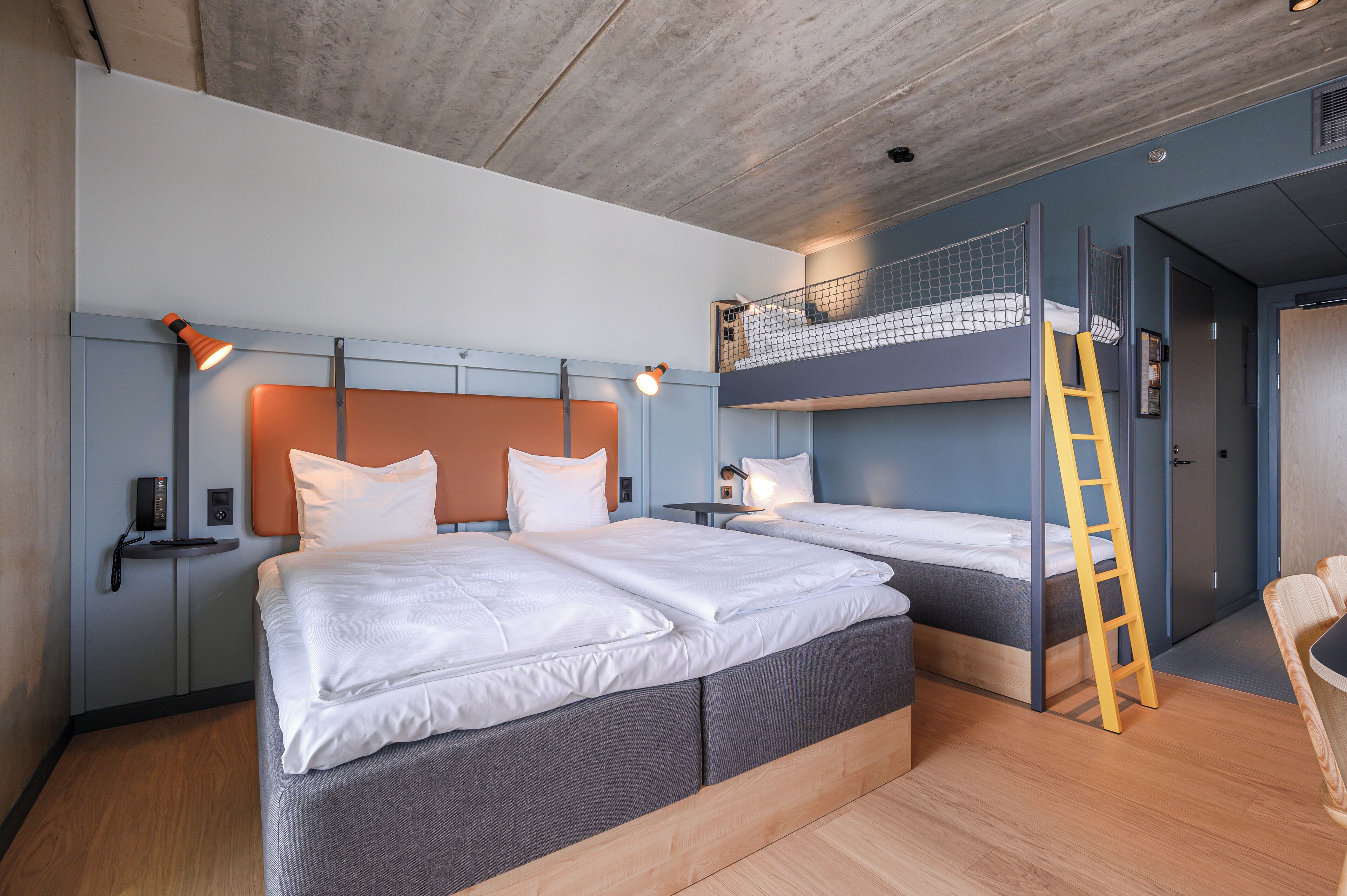 A modern hotel room with two beds, a bunk bed, a wooden ladder, and a small desk with a lamp.