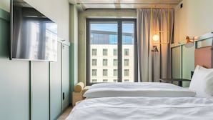 Une chambre d’hôtel avec un lit, un téléviseur fixé au mur, une fenêtre avec des rideaux et une vue sur un bâtiment à l’extérieur.