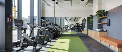 Un gimnasio con equipo de ejercicio, un sendero de césped artificial verde y una zona de recepción con un banco y plantas en macetas.