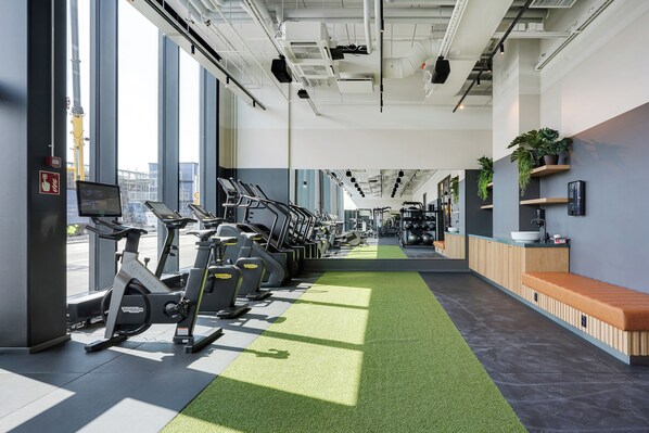 Une salle de sport équipée de matériel d’exercice, un chemin recouvert d’un gazon synthétique, et un espace d’accueil avec un banc et des plantes en pot.