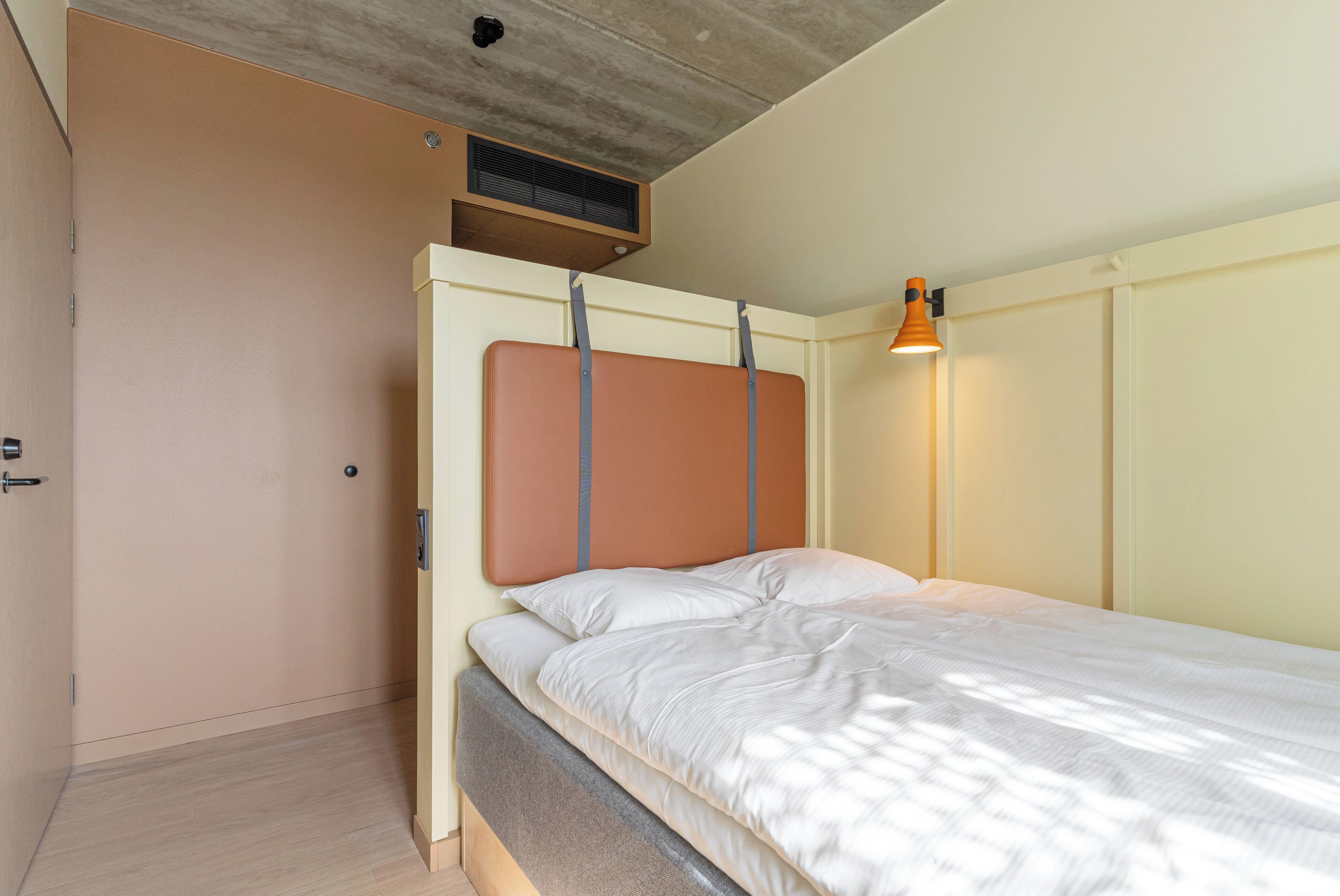 A bedroom with a bed, a headboard, a wall-mounted heater, and a bedside lamp.