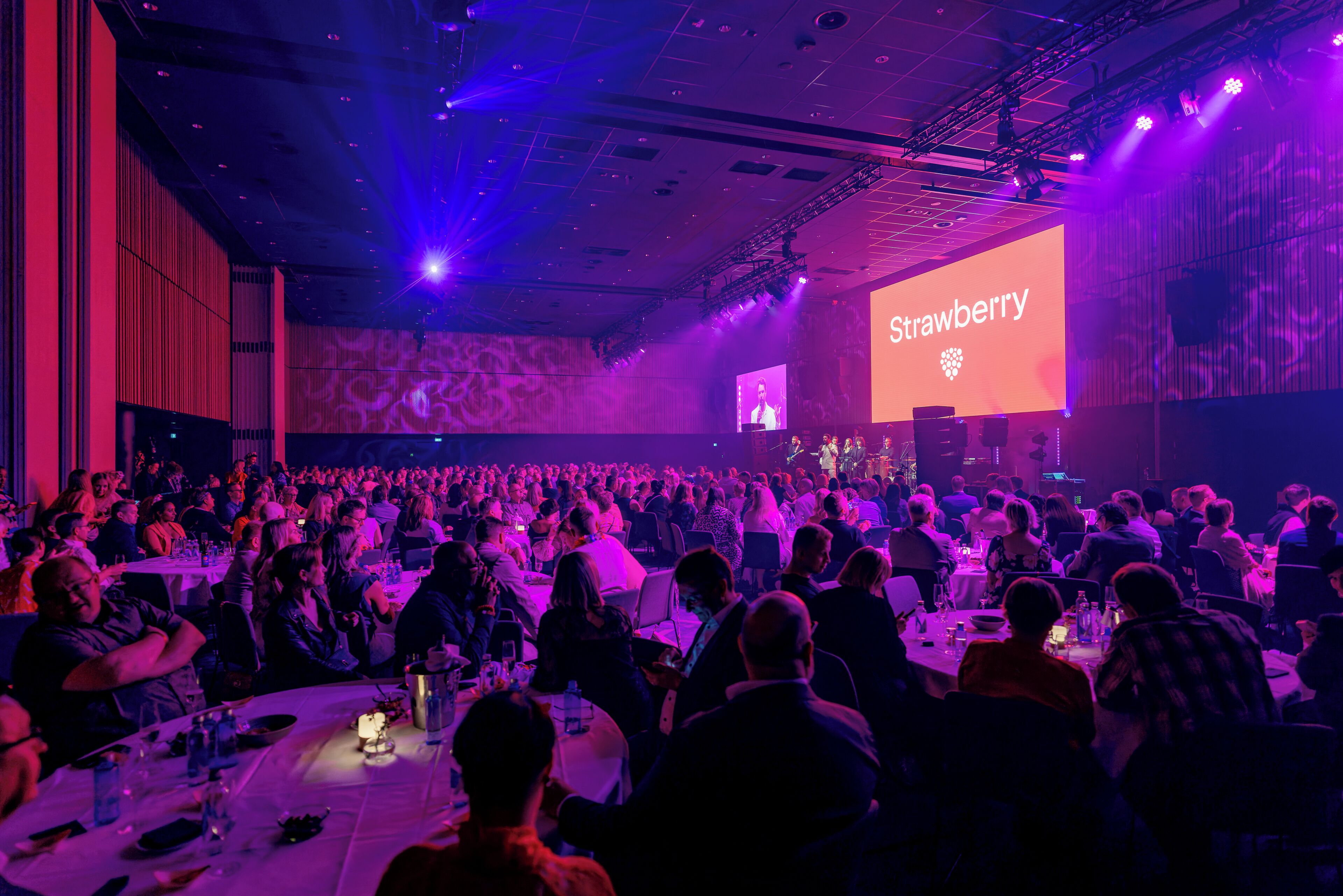 A conference room with a large audience seated at round tables, a stage with a presentation screen, and overhead lighting.