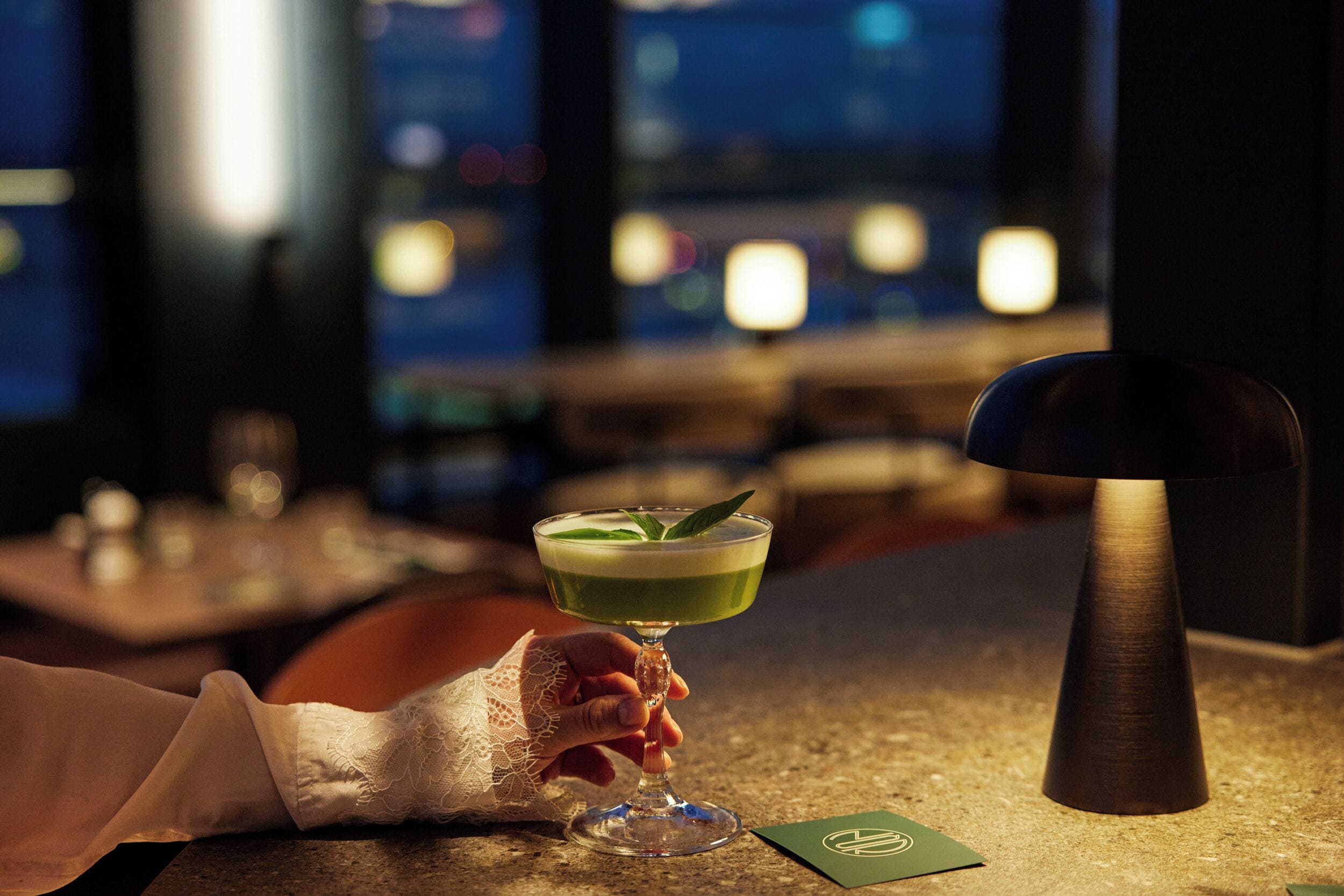 A cocktail in a glass with a green liquid and a garnish, held by a hand.