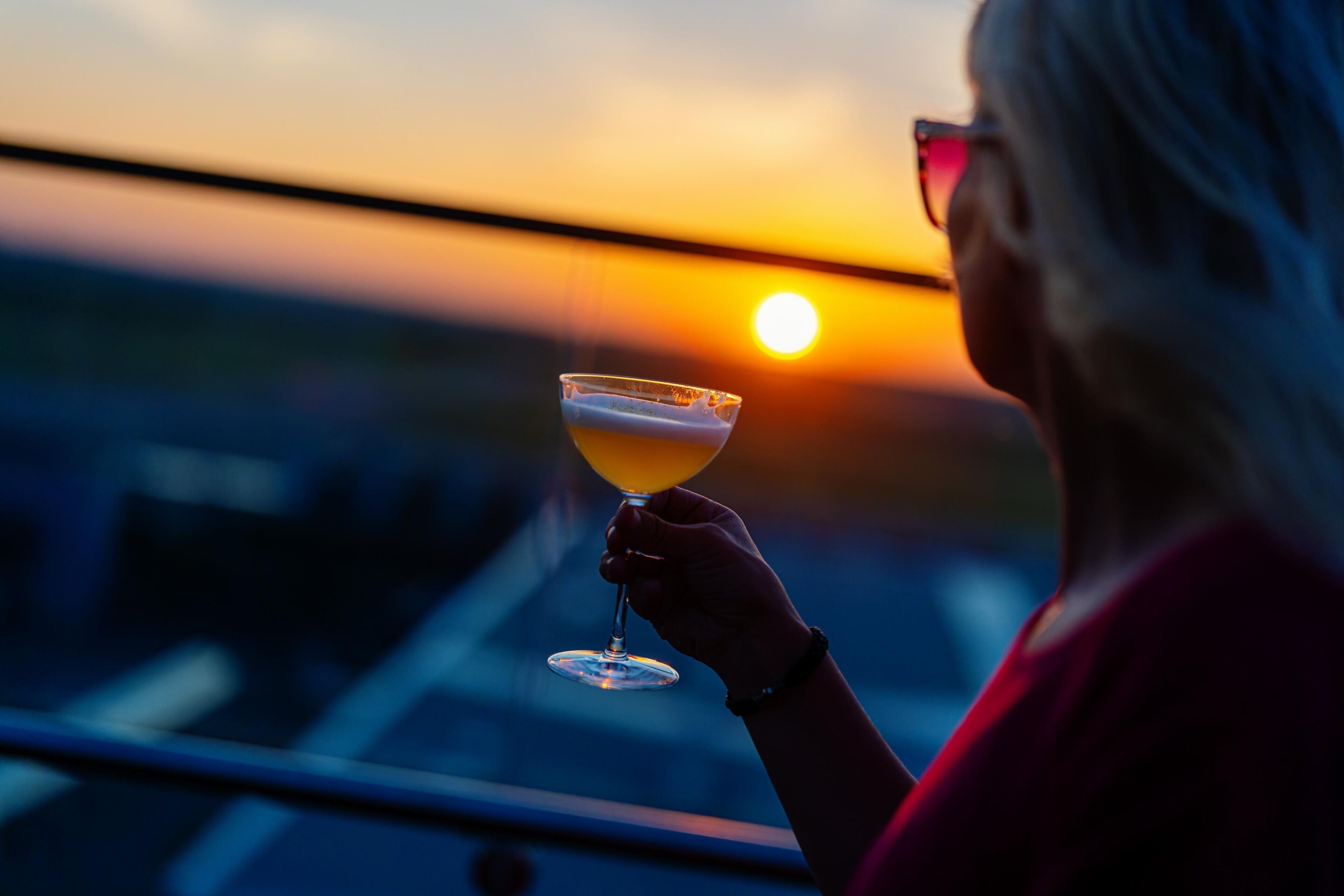 Henkilö, jolla on kädessään cocktail-lasi ja taustalla näkyy auringonlasku.