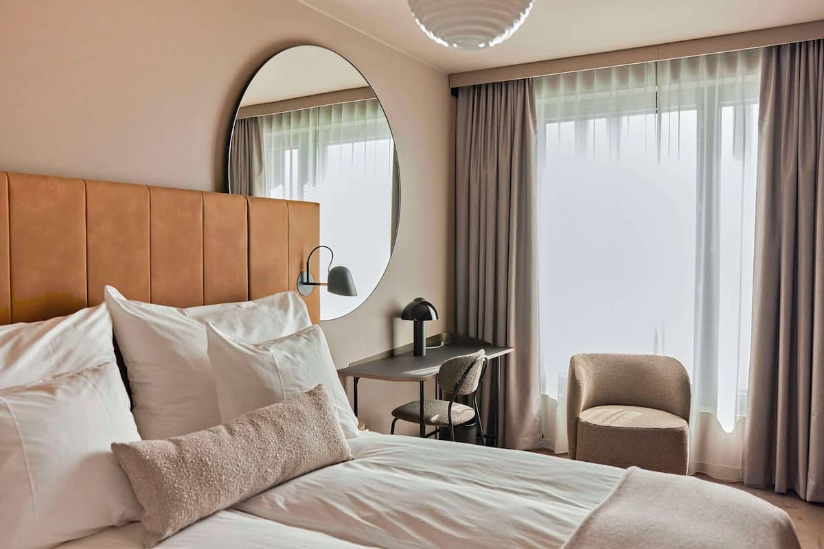 Ein Hotelzimmer mit einem großen Bett, einem Schreibtisch und einem Stuhl. An der Wand hängt ein runder Spiegel und es gibt ein Fenster mit Vorhängen.