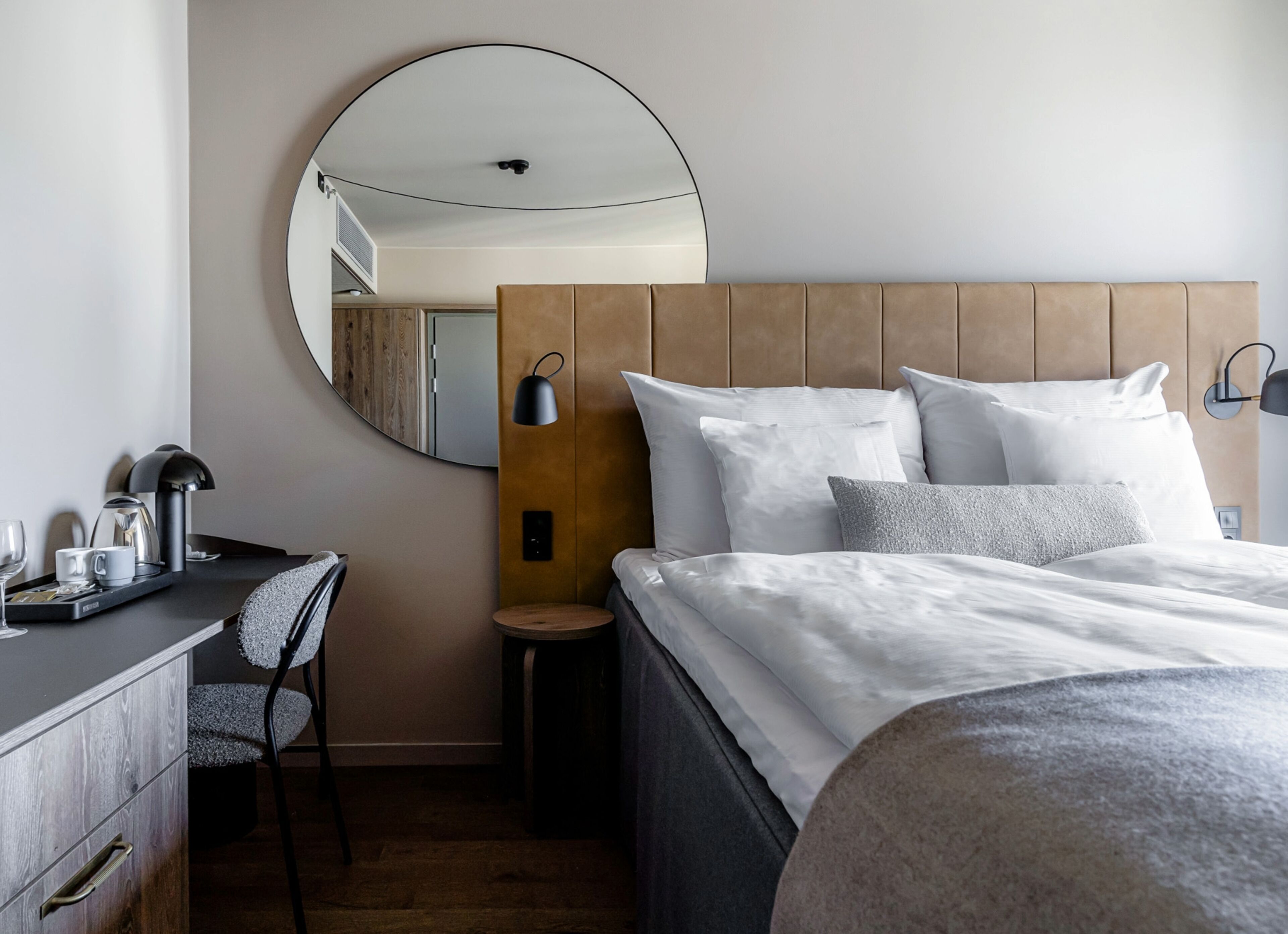 Una habitación de hotel con una cama, un escritorio, una silla, un espejo redondo y una mesita auxiliar.
