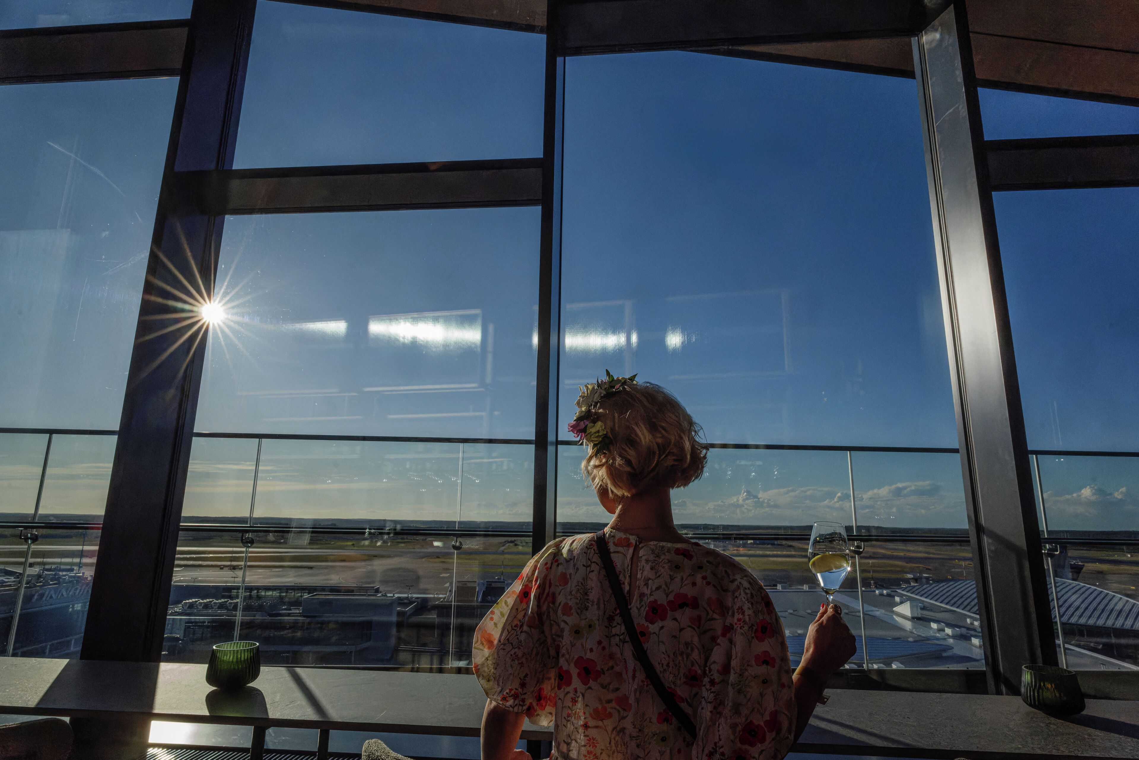 A person holding a wine glass, looking out from a high-rise building with large windows.
