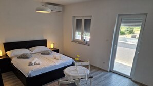 Un dormitorio con cama, mesitas de noche, una silla, una ventana con persianas y un equipo de aire acondicionado.