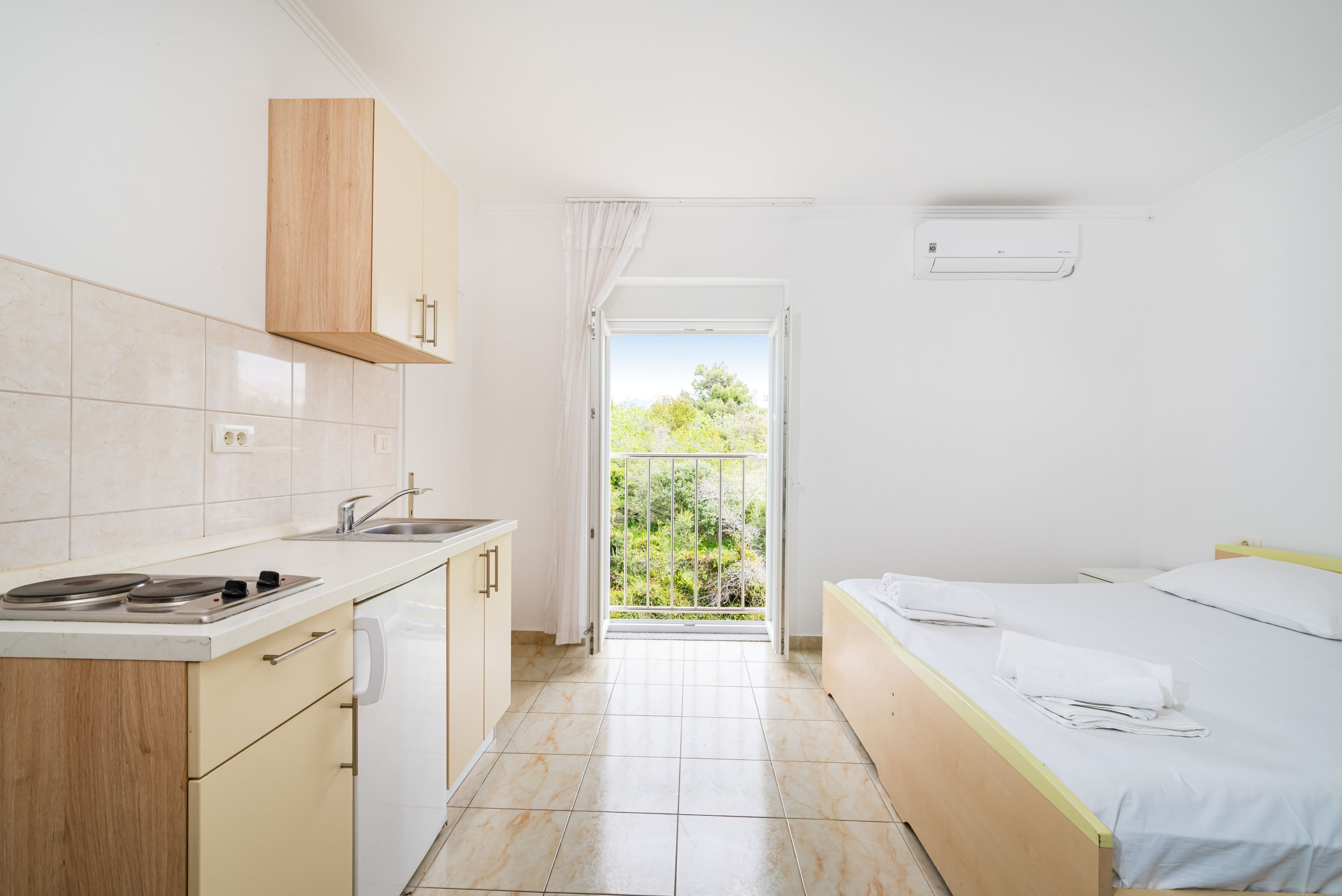A compact room with a kitchenette, a bed, and a balcony with a view of greenery.