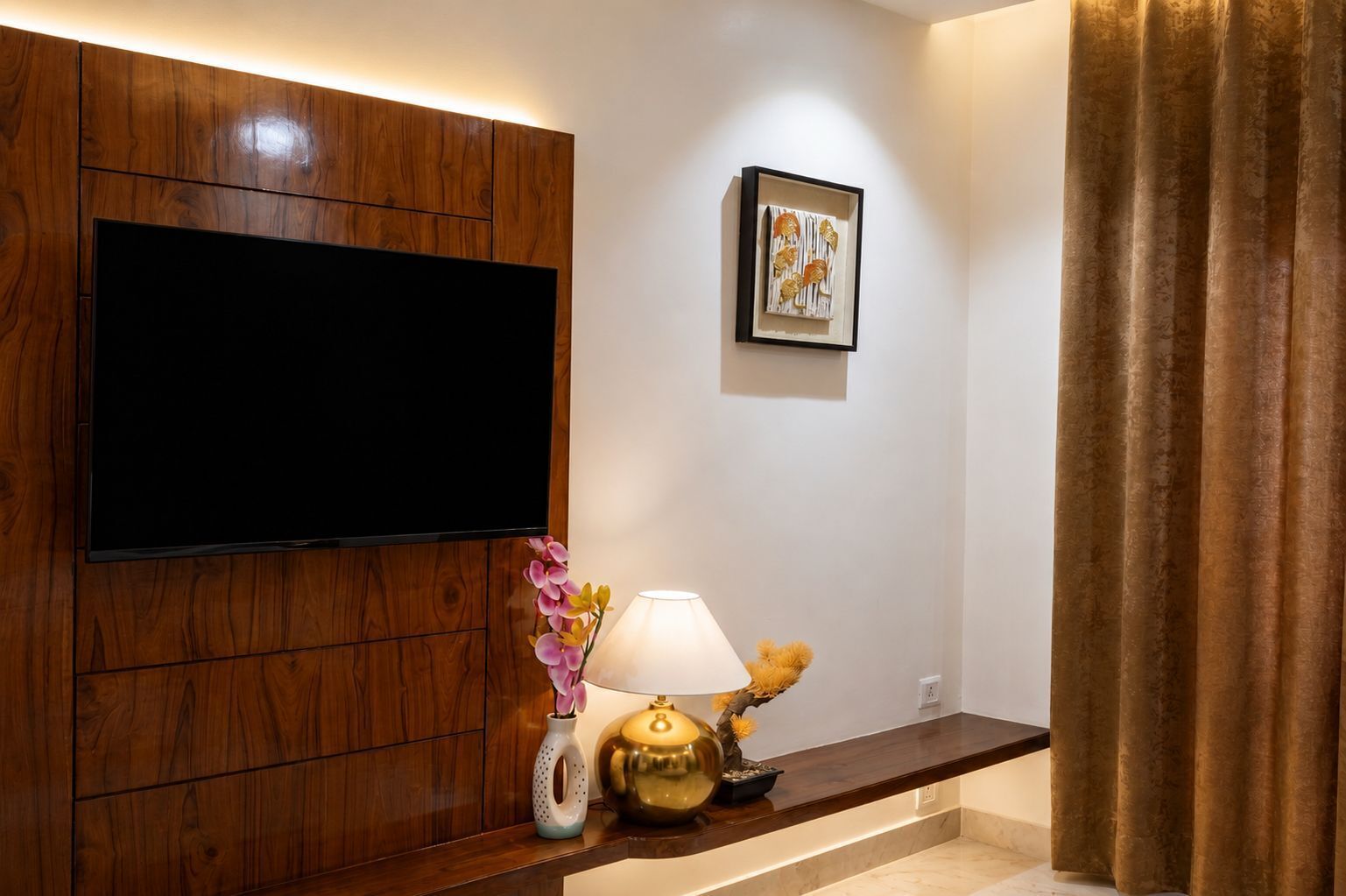 A modern room with a flat-screen TV mounted on a wooden panel wall, a small table with a lamp and decorative items, and a framed picture on the wall.