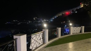 A nighttime view of a balcony with a tiled floor and metal railing, overlooking a cityscape with distant lights.