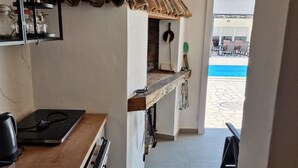 Une cuisine étroite au décor rustique, avec un mur en briques, des poutres en bois et une étagère intégrée où sont rangés des bocaux. On aperçoit une piscine à travers une porte ouverte.