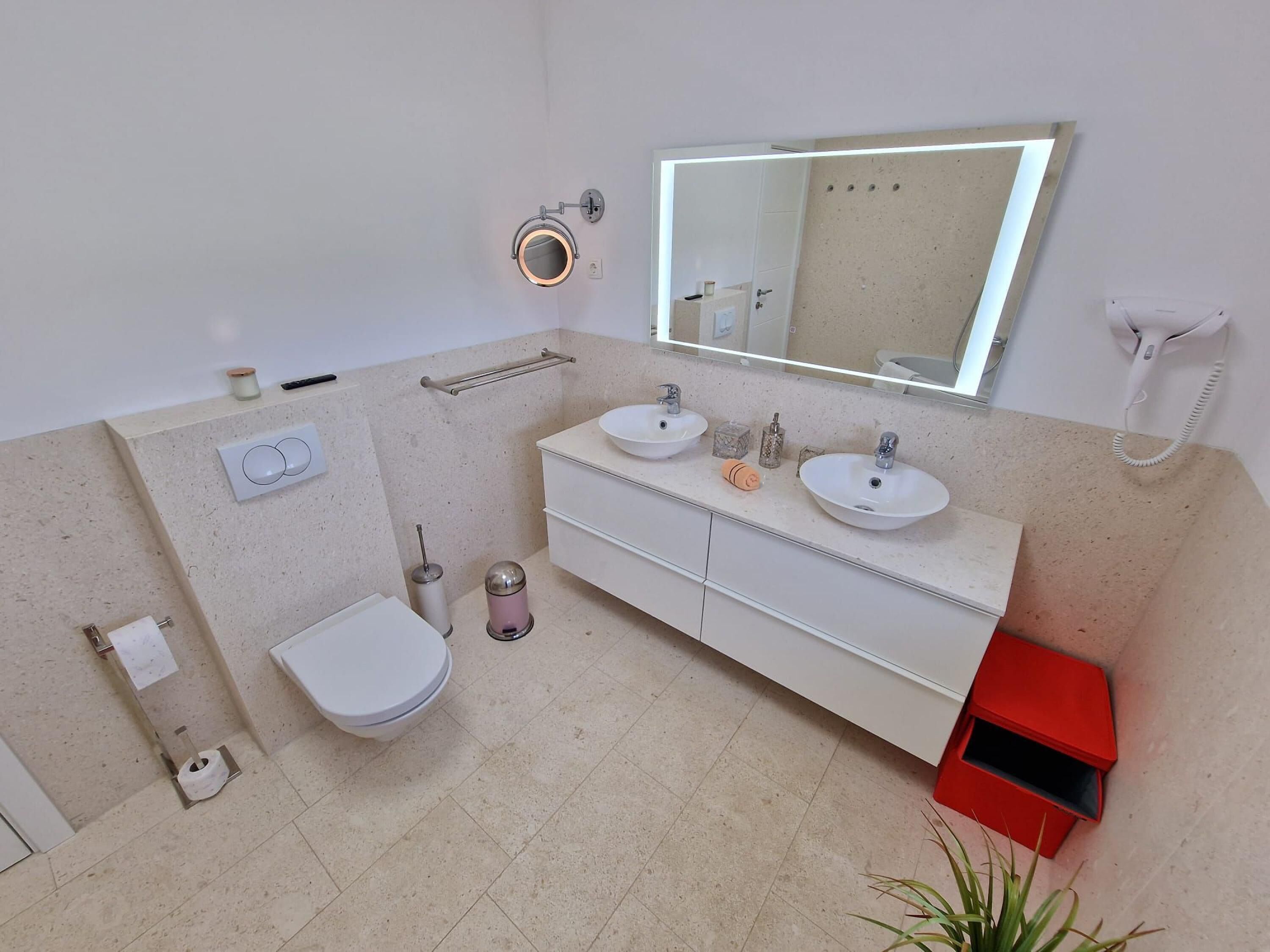 Un baño moderno con dos lavabos, inodoro, bidé, espejo con iluminación integrada, teléfono y un caja roja para guardar objetos.
