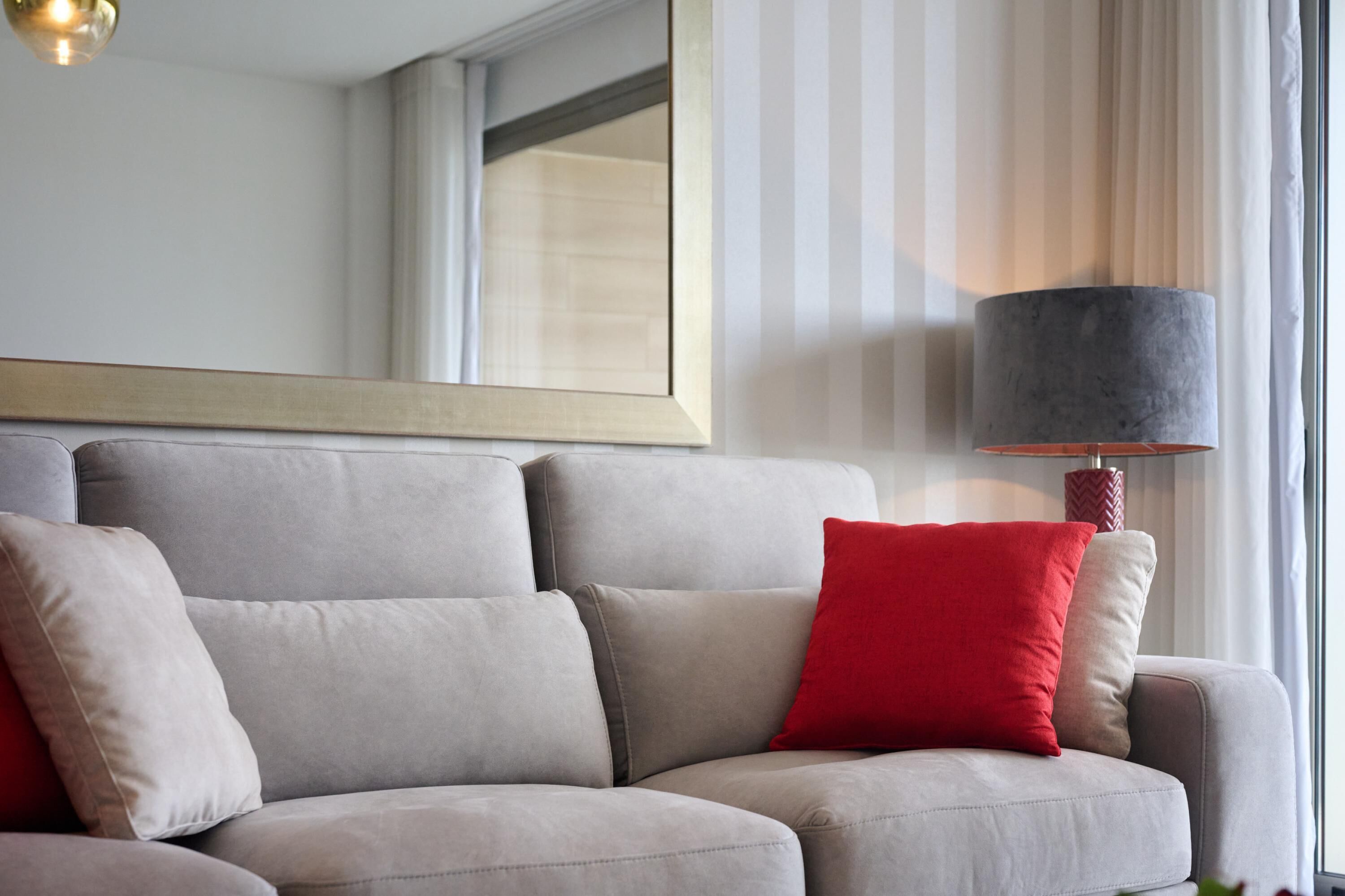 A modern living room with a grey sofa, red and beige cushions, a framed artwork above the sofa, and a lamp with a grey shade.