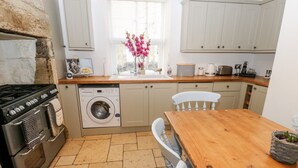 A kitchen with a stone wall, a washing machine, a stove, a wooden table, and a window with flowers. - The Cottage (Leeds)