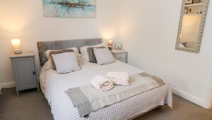 A neatly made bed with a textured blanket and pillows, a bedside table with a lamp, and a painting above the bed. - The Cottage (Leeds)
