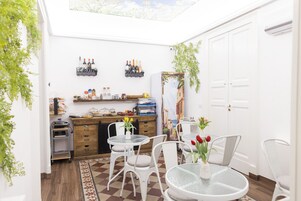 Una cocina compacta con armarios blancos, encimera de madera y un estante empotrado para vinos. Hay dos sillas blancas alrededor de una mesita redonda, y se ve una planta en maceta.
