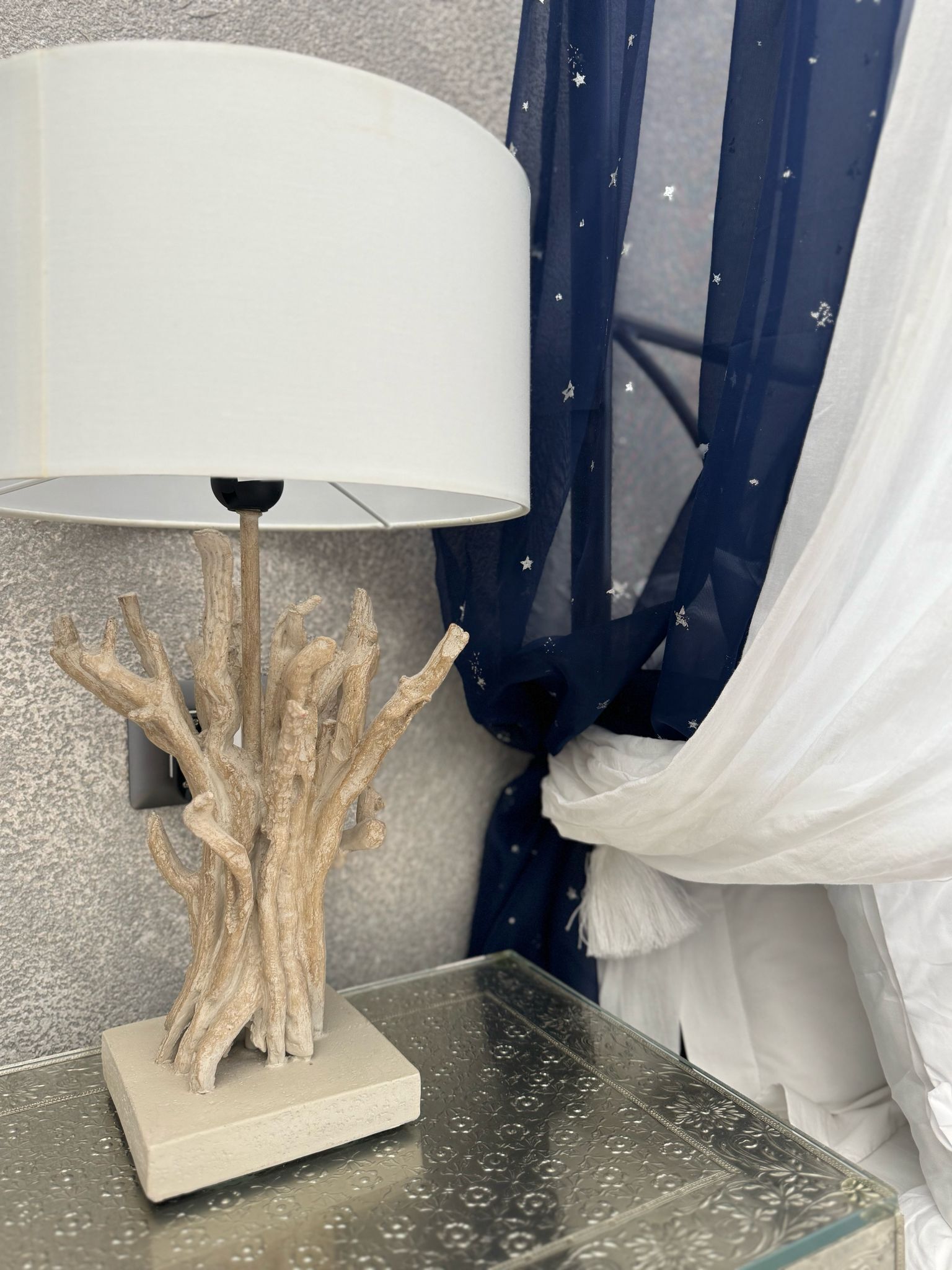Eine Lampe mit weißem Lampenschirm auf einem Glastisch, ein dekorativer Baumstamm und ein blauer Vorhang mit Sternen.