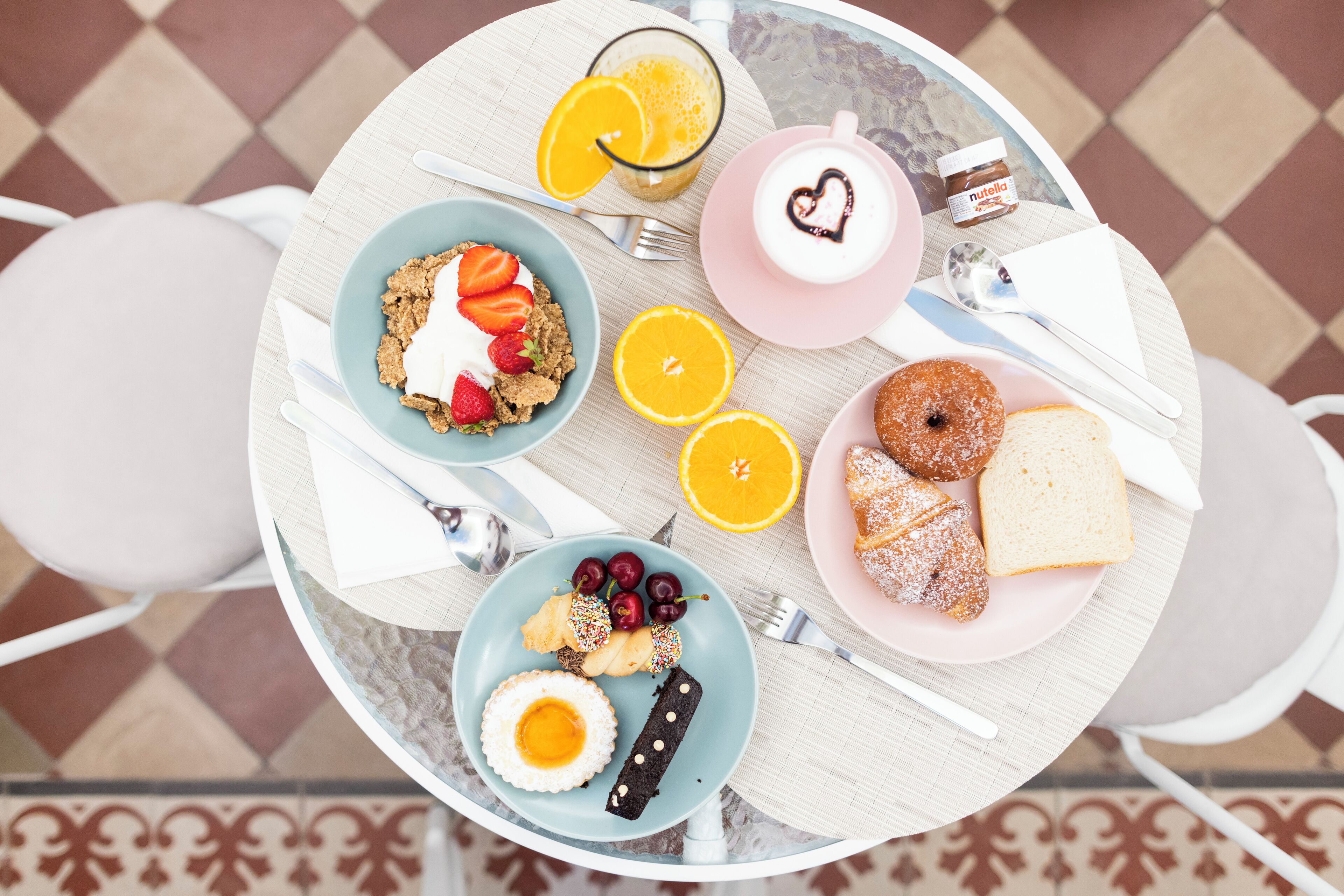 Un desayuno con diversos alimentos sobre una mesa redonda, incluyendo un plato con fresas, un vaso de zumo de naranja, un plato con rosquillas y una taza de café con diseño de corazón.