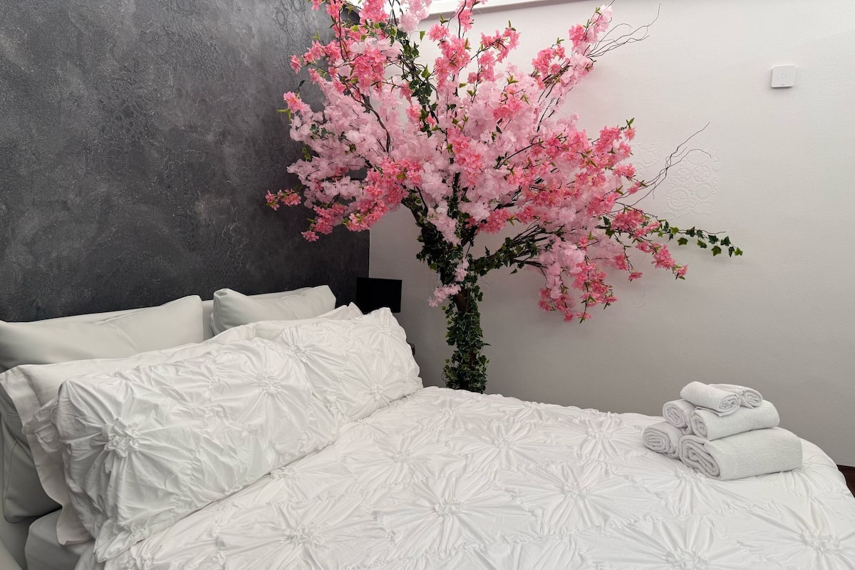 Ein Schlafzimmer mit einem großen Bett, weißer Bettwäsche, einem an der Wand montierten Thermostat und einer blühenden rosa Pflanze.