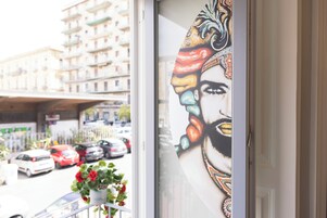 Una ventana con vista a una calle, un edificio con balcones y una planta en maceta con flores rojas.