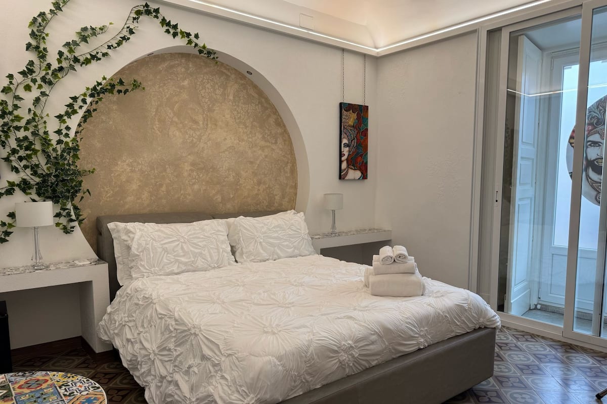 Ein modernes Schlafzimmer mit einem großen Bett, einem dekorativen Wandbogen, einem Gemälde und Blick durch eine Glastür.