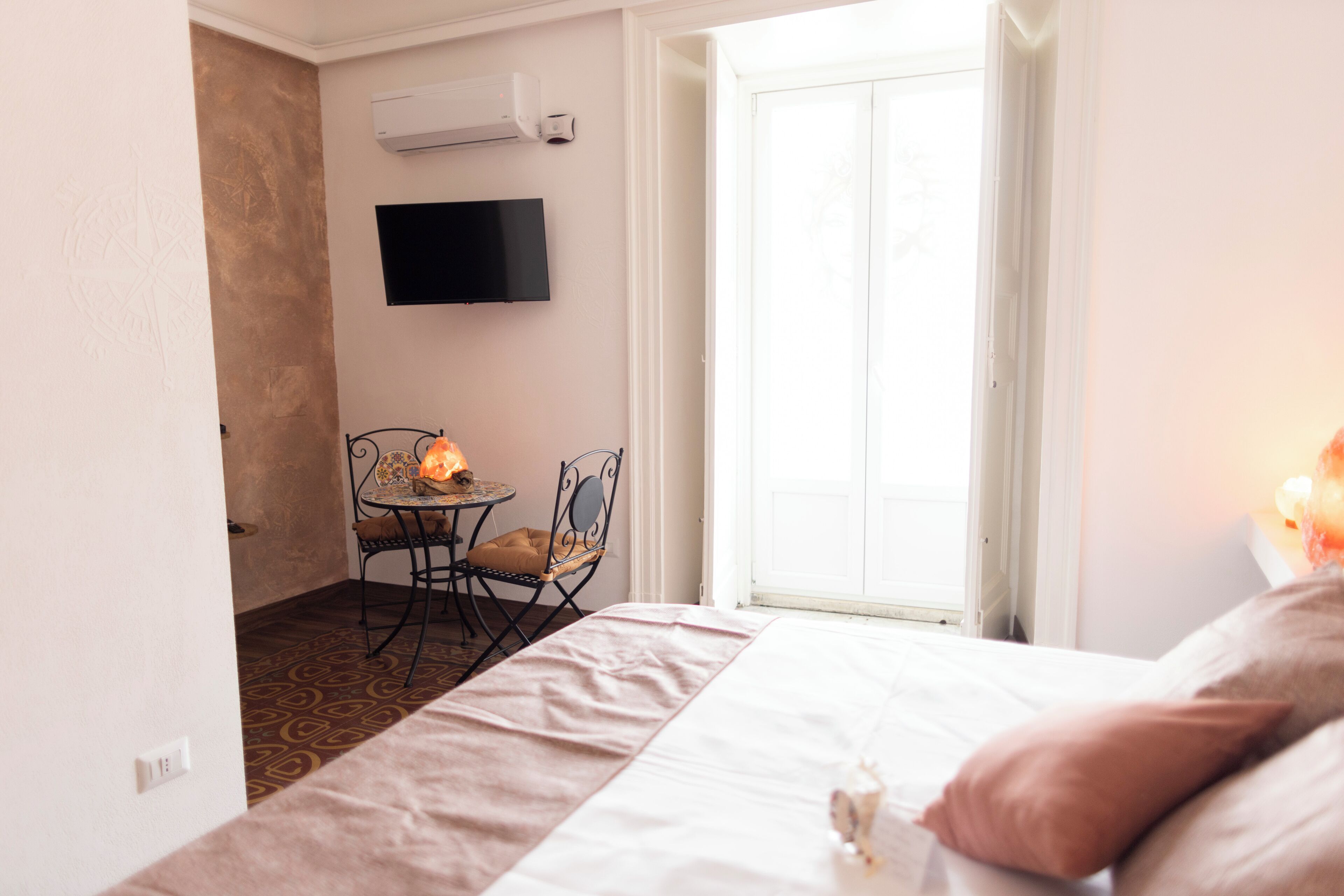 A hotel room with a bed, a small table with a chair, a flat-screen TV mounted on the wall, and a sliding glass door leading to a balcony.