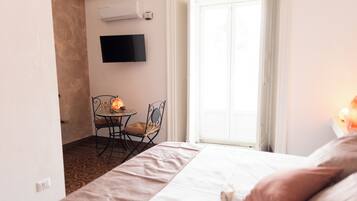 A hotel room with a bed, a small table with a chair, a flat-screen TV mounted on the wall, and a sliding glass door leading to a balcony.