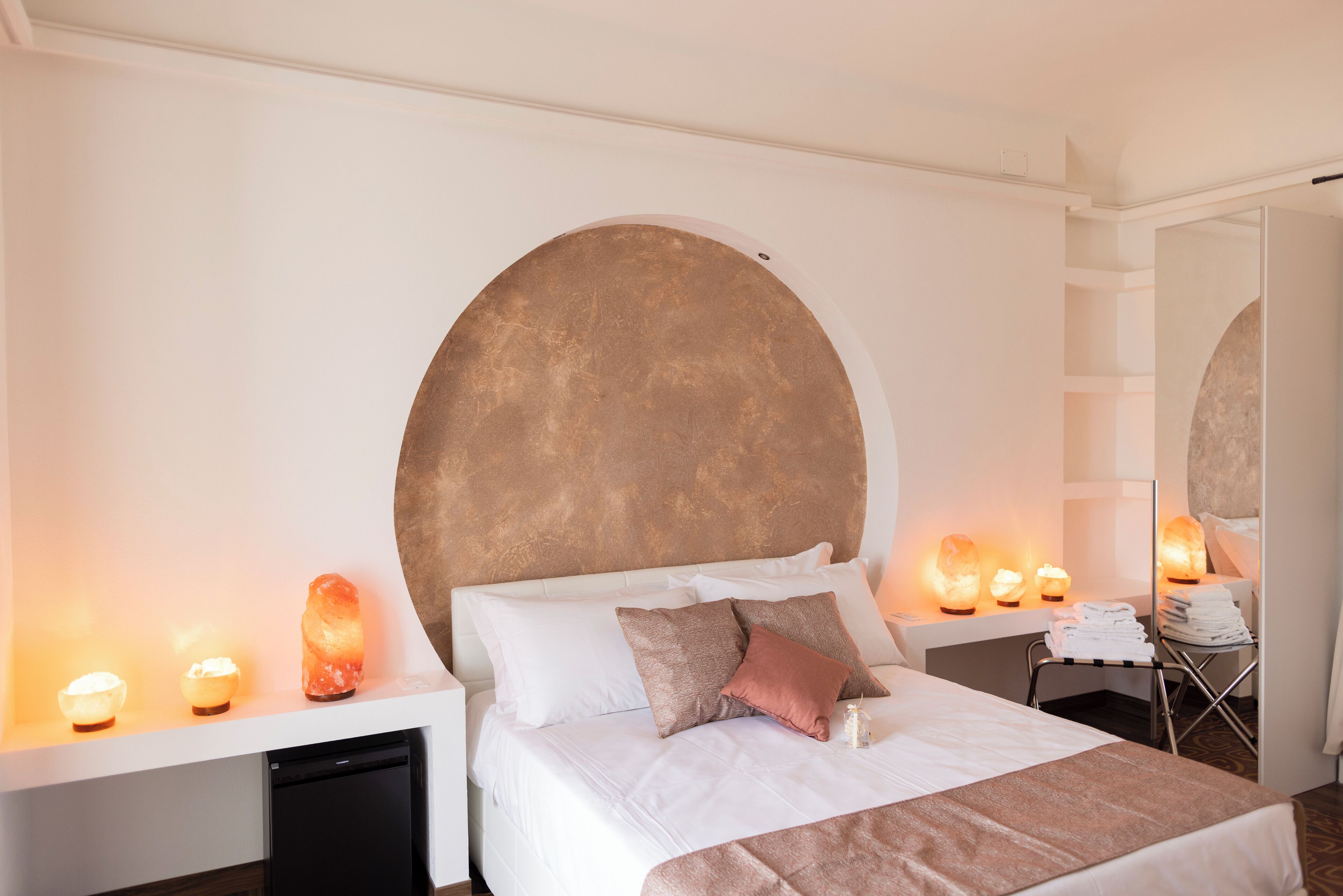 Ett modernt hotellrum med en stor säng, vita sängkläder, en rund väggdekoration och flera saltlampor på nattduksborden.