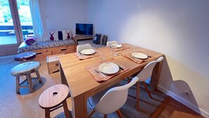 Zona pranzo con tavolo in legno apparecchiata per quattro persone, una poltrona con cuscini, una televisione e vista sull'esterno.