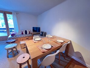 A dining area with a wooden table set for four, a sofa with cushions, a television, and a view of the outdoors.