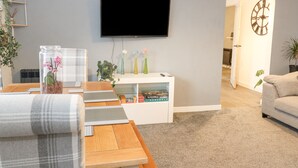 A modern living room with a flat-screen TV mounted on the wall, a coffee table with a remote control, and a bookshelf with decorative items. - 22 Moray Place (Fort William)