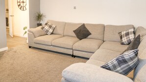 A living room with a beige sectional sofa, a clock on the wall, and a plant. - 22 Moray Place (Fort William)