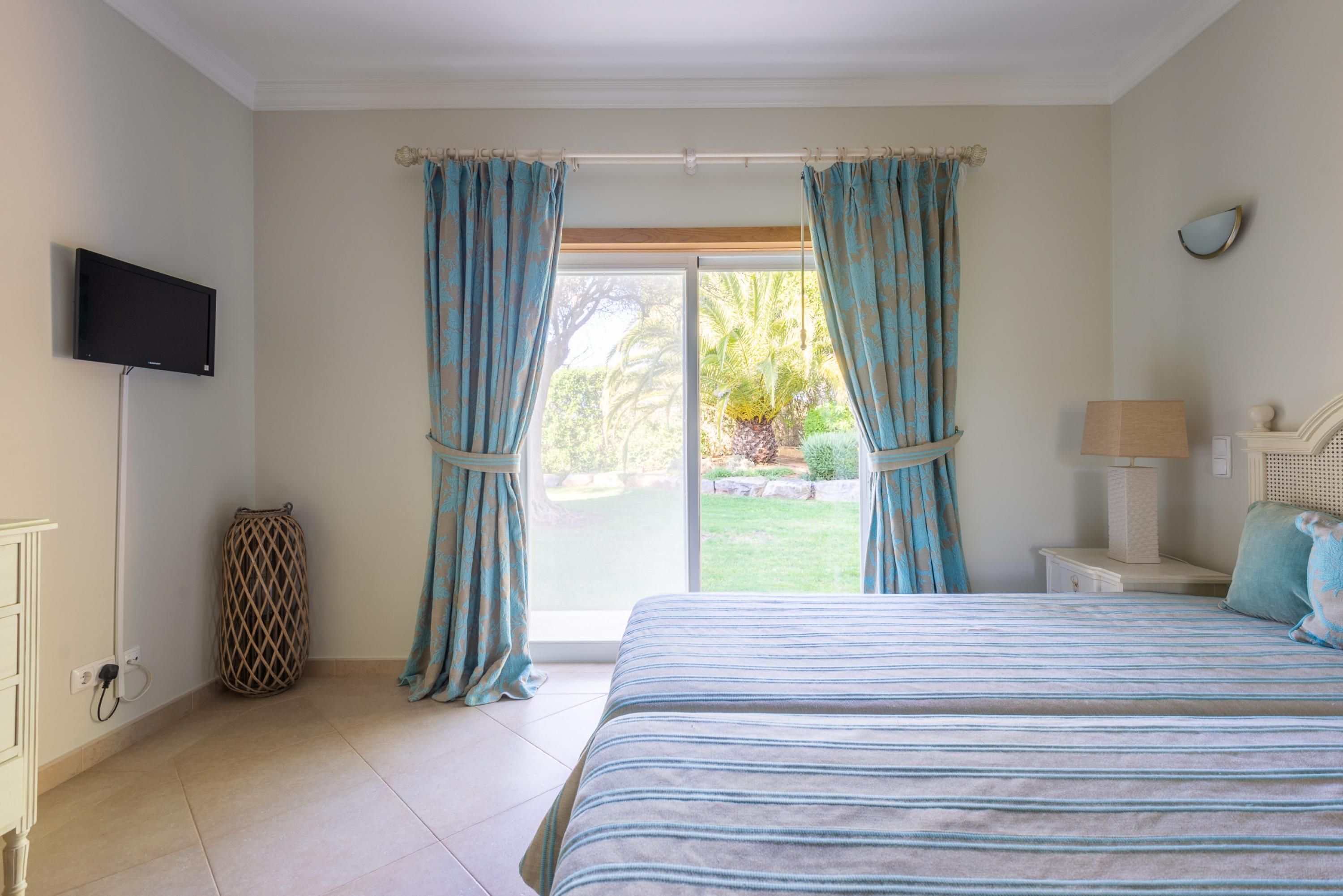 A bedroom with a bed, curtains, a television, a bedside table, and a view of the outdoors.