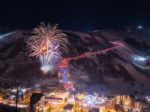 Un pueblo con una montaña destacada al fondo, un sendero iluminado y un gran espectáculo de fuegos artificiales.