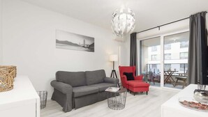 Un salon moderne avec un canapé gris, un fauteuil rouge, une table basse blanche et une grande fenêtre donnant sur des immeubles.